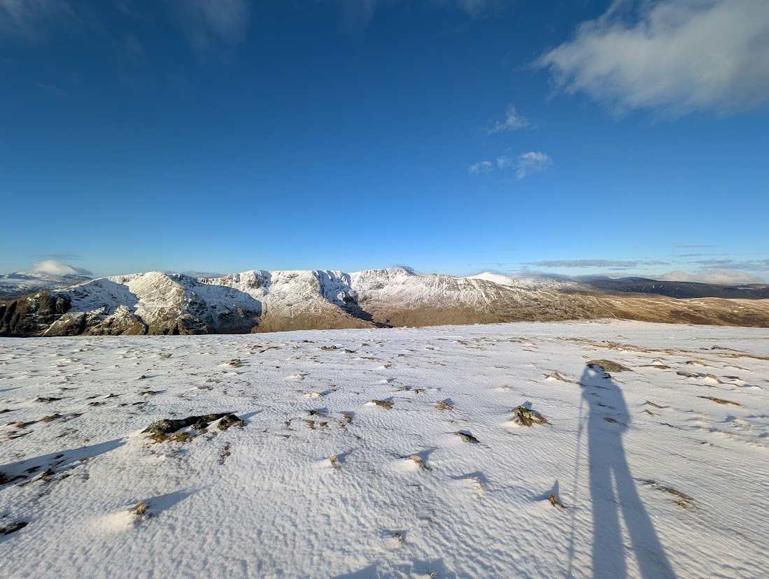 Snow lying across a high Lakeland summit under a deep blue winter sky, with the snow-covered Helvellyn range beyond and the photographer's long shadow across the snow.