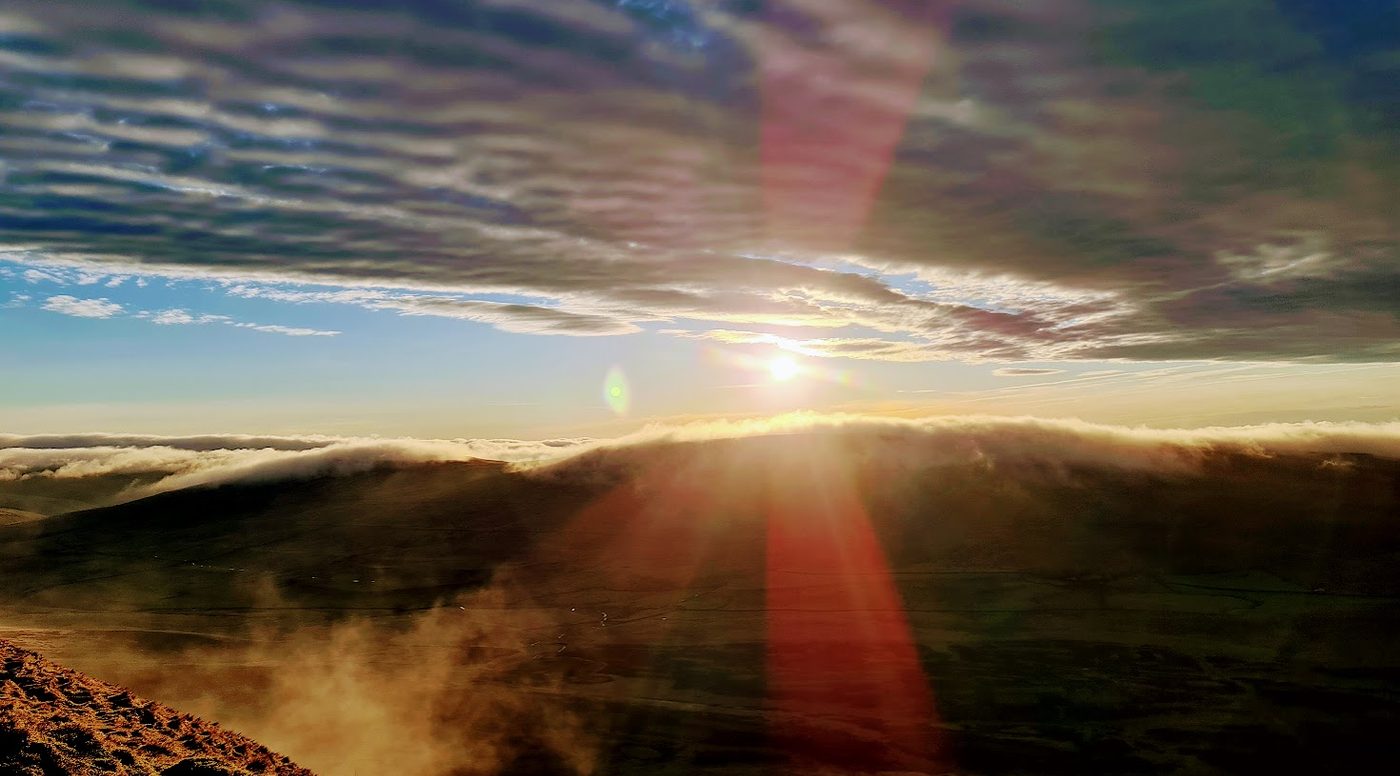 Sunrise seen from a Lakeland summit standing above a temperature inversion, with a sea of cloud filling the valley below and the sun low over a distant ridge.