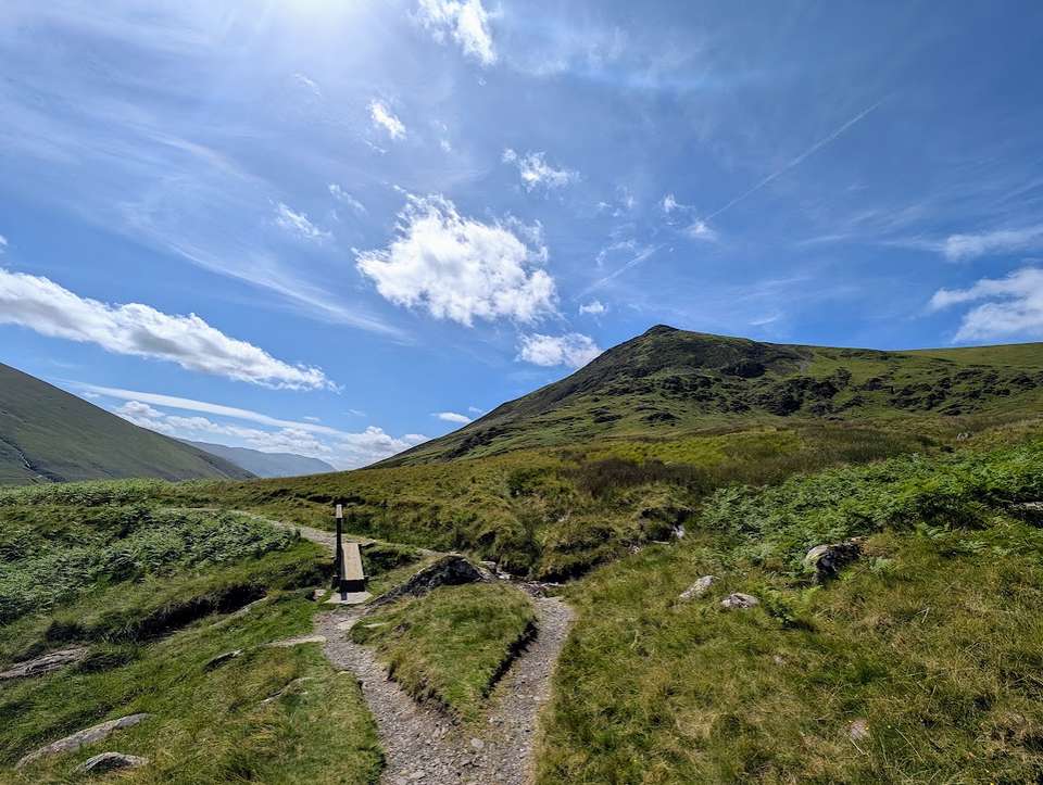 A stony path forking beside a wooden signpost in a sunlit Lakeland valley, a steep green fell rising ahead under blue sky.