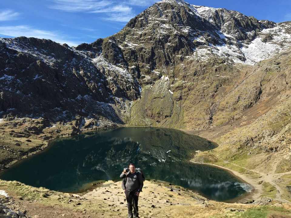 A walker stands by a mountain tarn below the snow-streaked summit of Snowdon under a clear blue winter sky.