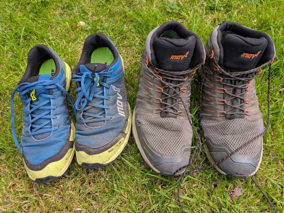 Two pairs of footwear side by side on grass: blue-and-yellow low trail-running shoes on the left, and grey-and-orange mid-height walking boots on the right.