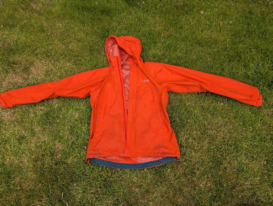 A bright orange Rab waterproof shell jacket laid out flat on grass, a high-visibility colour that stands out clearly against the green.