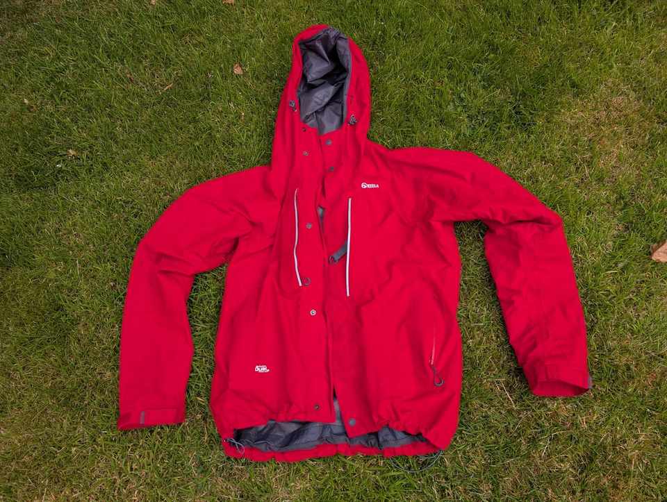 A red waterproof hard-shell jacket with a hood and taped seams laid out flat on grass.