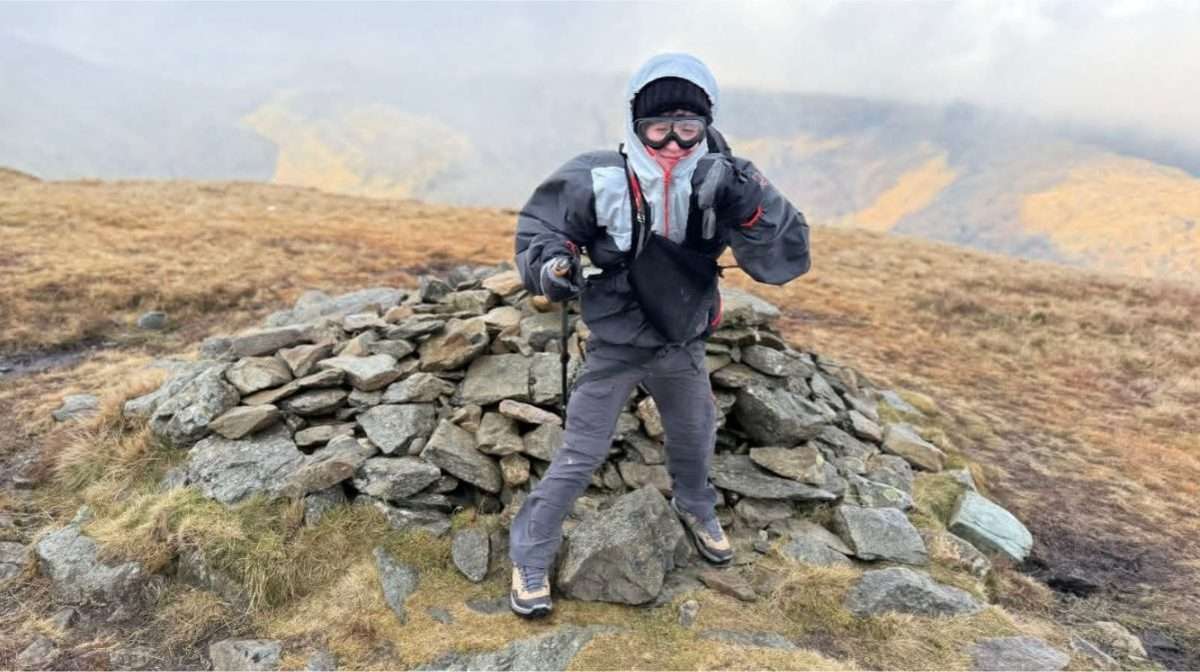 A walker leaning into strong wind on an exposed Lake District ridge, jacket and gear buffeted, cloud streaming past.