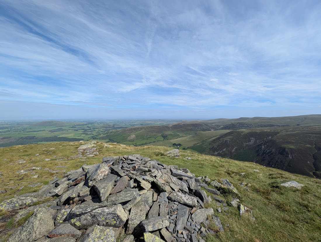 Bakestall - a stony summit cairn on grass, green lowland plain stretching beyond and a steep fellside falling away to the right