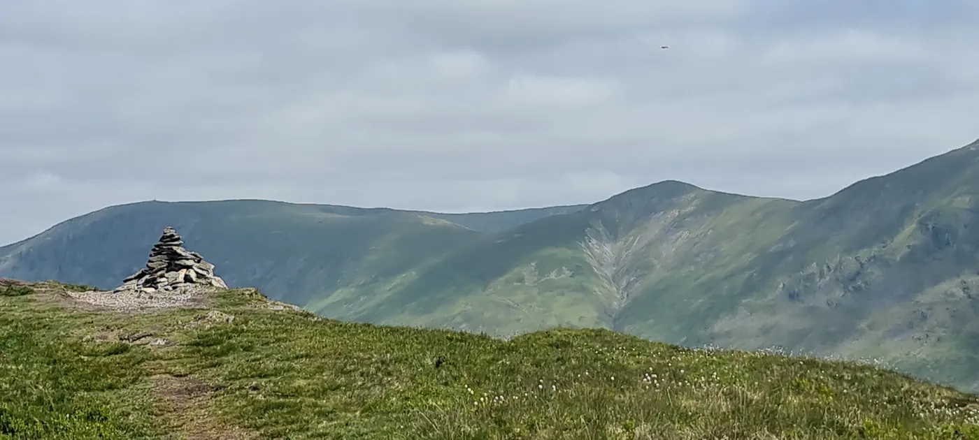 Baystones - a tall conical cairn on a grassy ridge, green fells with a steep gully opposite under overcast sky