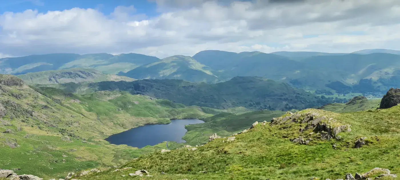 Blea Rigg - a dark mountain tarn cradled in green fellsides, rolling summits receding to the horizon under summer cloud