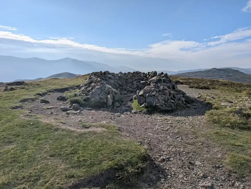 Bleaberry Fell - a big stone summit cairn on a stony top, hazy layered fells fading into the distance beyond