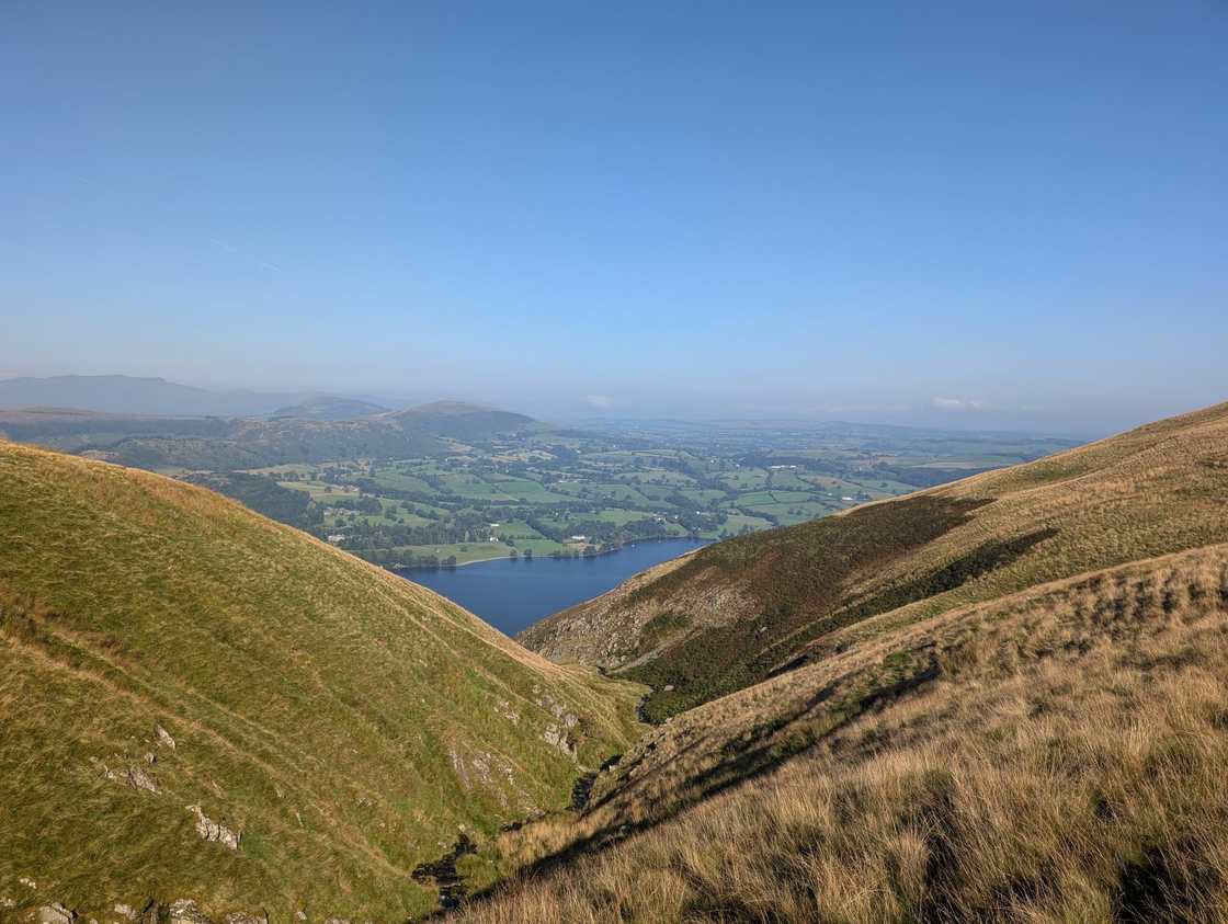 Bonscale Pike - grassy fellsides framing a steep combe that drops to Ullswater, with green fields and haze beyond