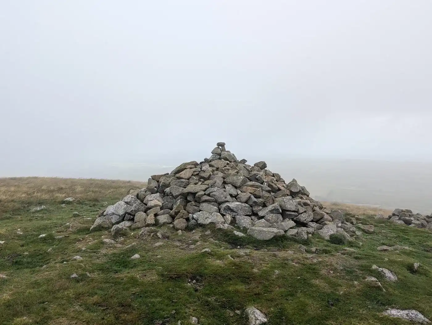 Brae Fell - a neatly built conical stone cairn on a grassy summit, distant low ground lost in grey haze