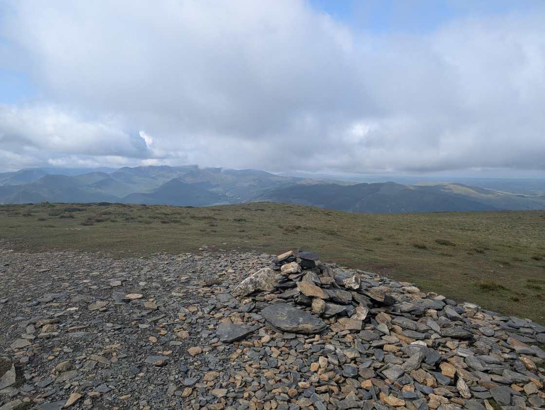 Carl Side - a low slate cairn on a flat stony summit, a long line of distant fells under cloudy sky