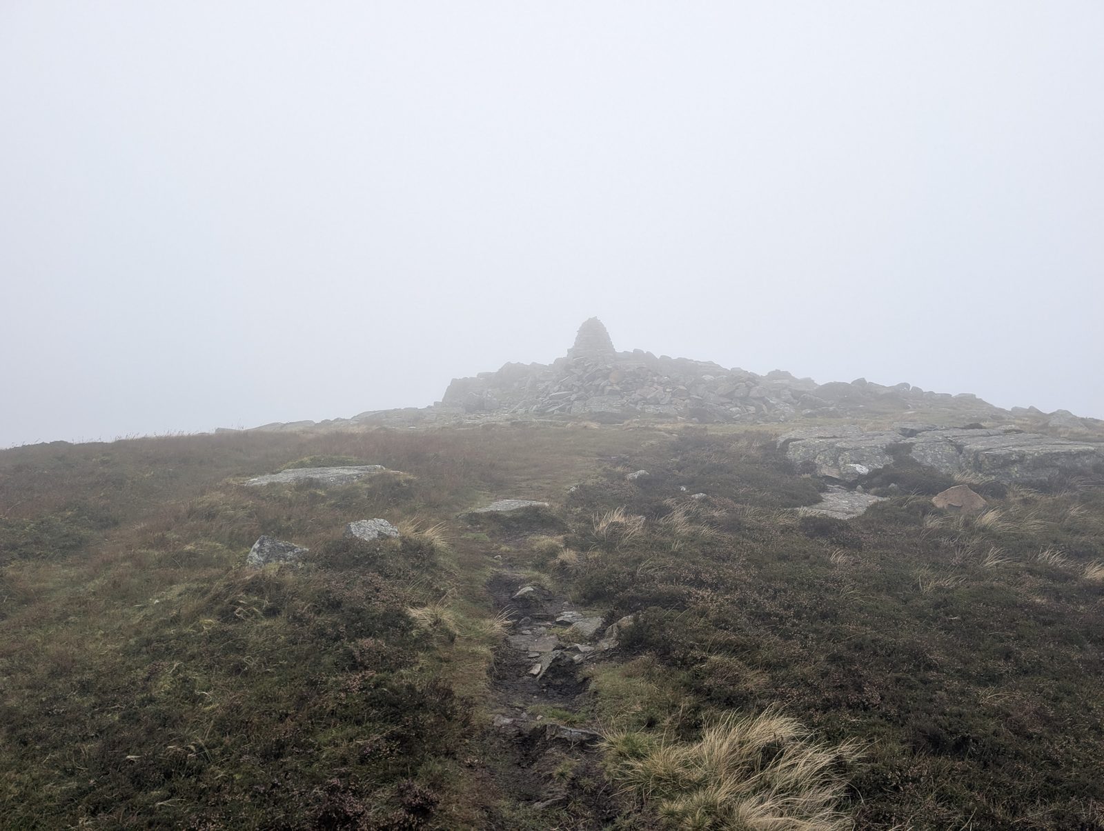 A worn path through heather climbs to the rocky crown of Carrock Fell, its tall summit cairn on the skyline in thick mist.