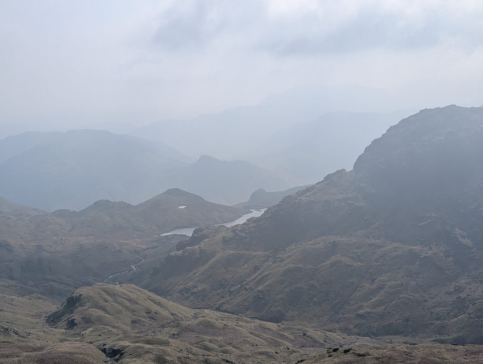 Codale Head - looking down from the summit slopes to a tarn in a hazy valley, a rocky knoll rising to the right and ridge after ridge fading into mist