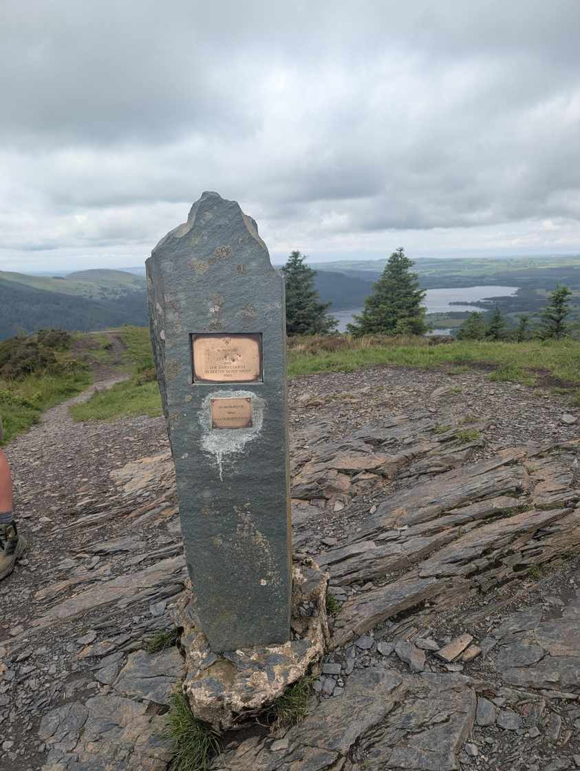 Dodd - a tall slate summit pillar with a bronze plaque, conifers and a distant lake beyond under grey cloud