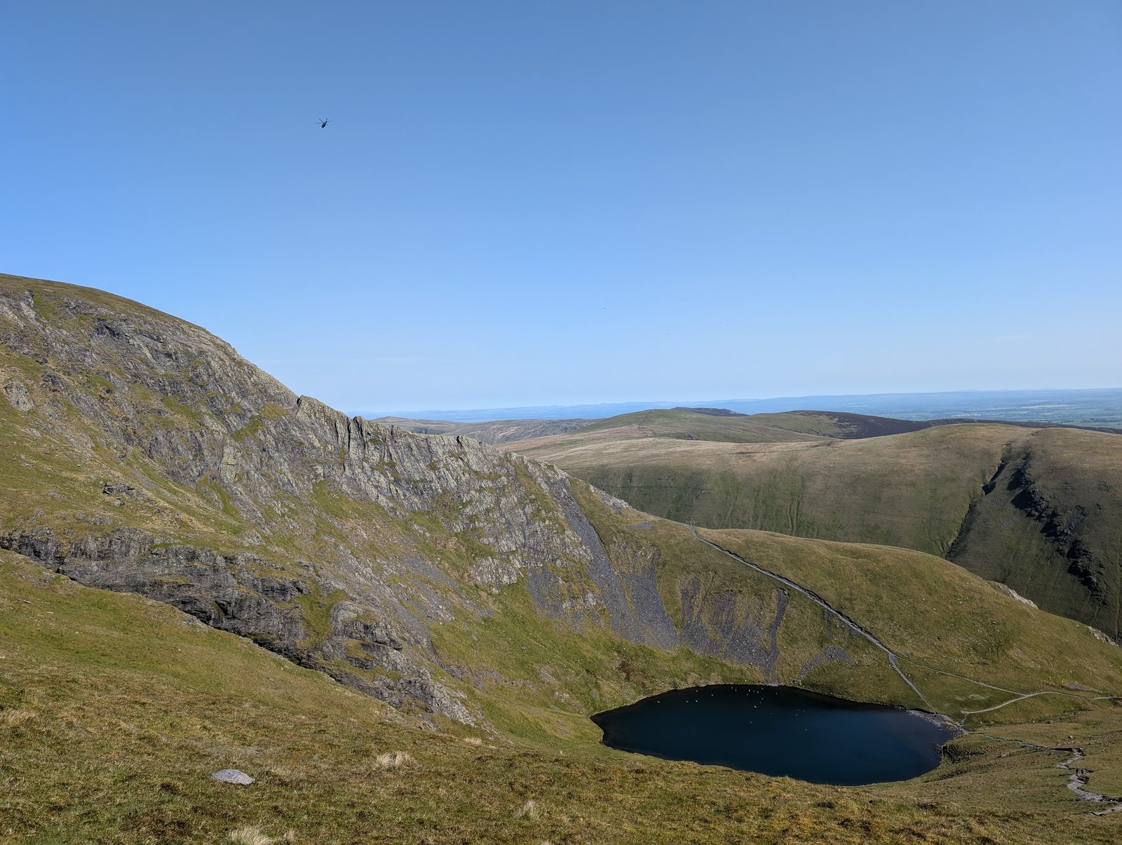 A dark tarn below a wall of crags and a sharp ridge, seen from the grassy flank of the fell