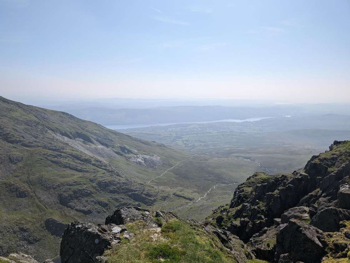 Dow Crag - dark crags above a green valley falling to Coniston Water, hazy estuary and lowlands far beyond