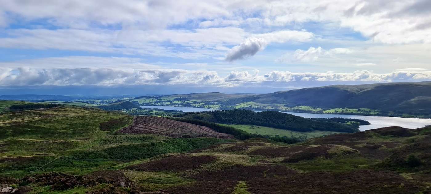 Heather-clad slopes of Gowbarrow falling away to Ullswater, the lake winding between rolling fells under a bright, cloud-dappled sky