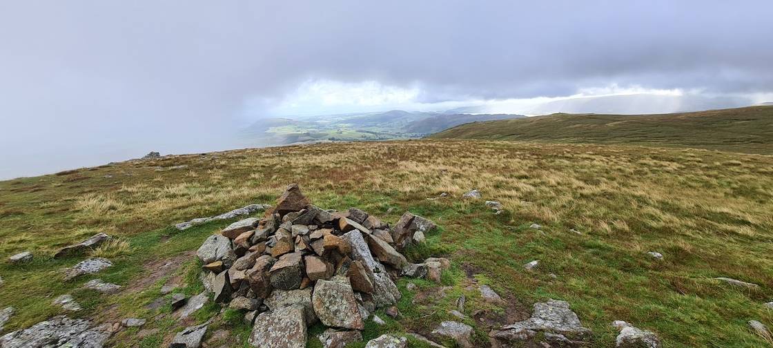 Hart Side - a stone cairn on a broad grassy summit, distant fells and lowlands beneath showery grey cloud