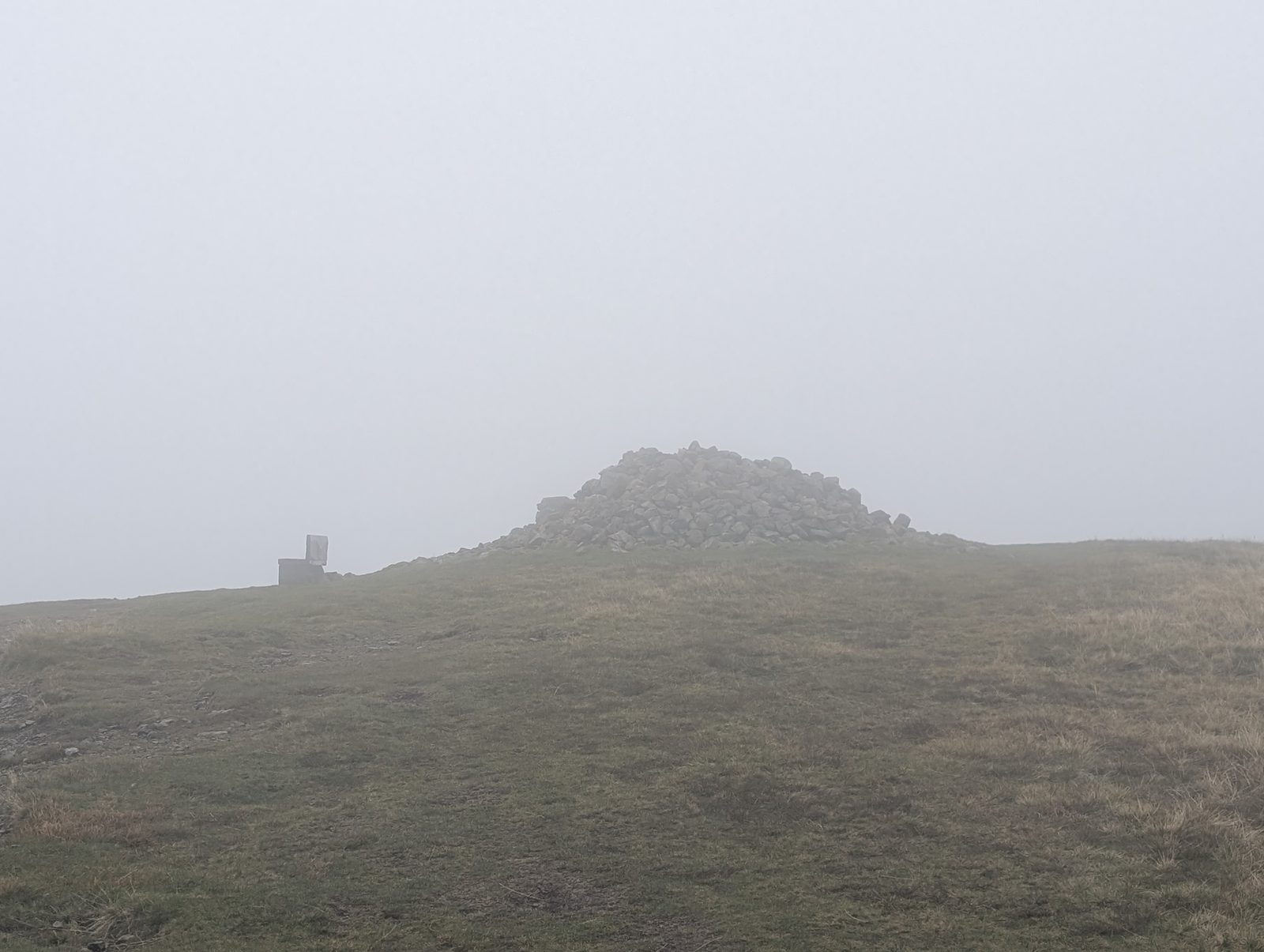 The big stone cairn on High Pike's grassy summit in thick mist, with a stone seat to its left.