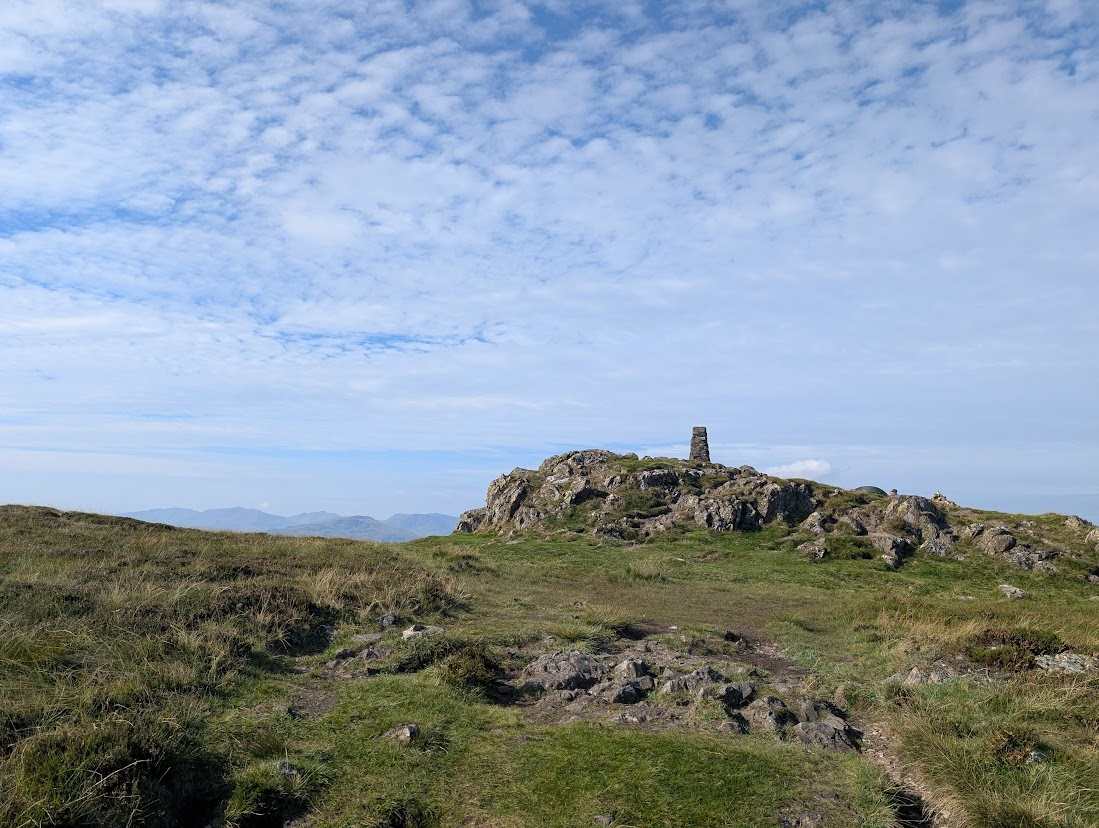 High Seat - a trig pillar on a rocky outcrop above grassy moorland, distant fells low on the horizon under blue sky