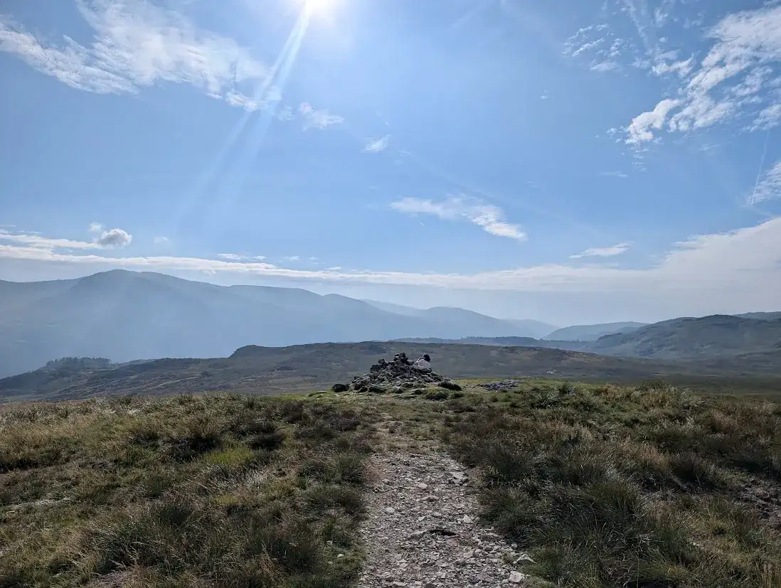 High Tove - a path crossing grassy moorland to a low cairn, hazy fells beyond and the sun flaring overhead