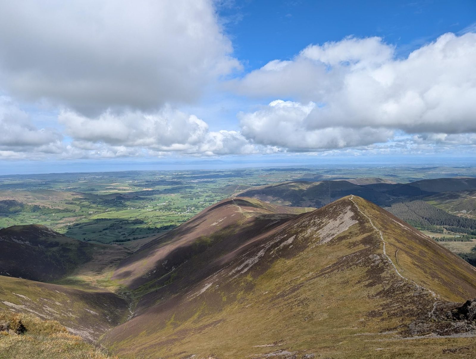 Looking down a heather ridge to a pointed grassy top with a path along its crest, a second rounded ridge beside it, and green farmland running out to the coast under big white clouds