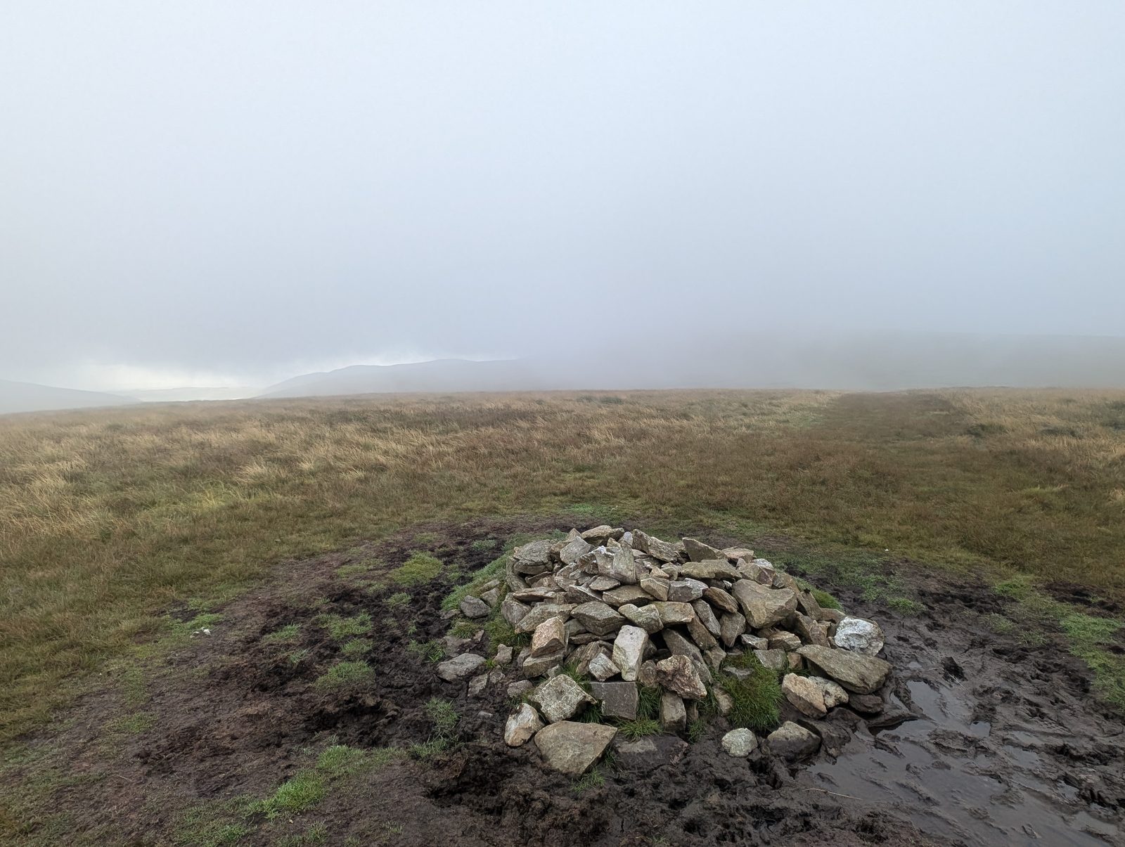 A modest cairn on a grassy top, with the ridge fading into mist beyond