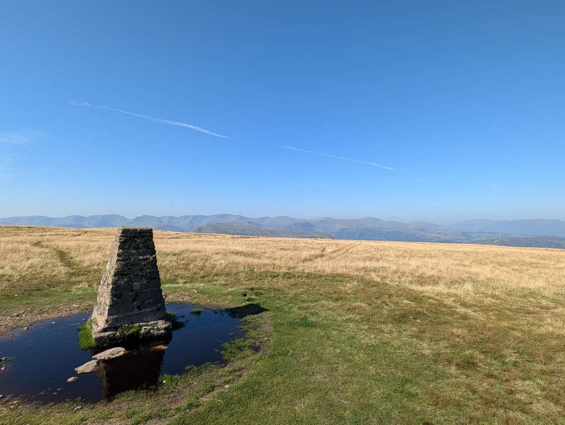 Loadpot Hill - a trig pillar ringed by a small pool on a broad grassy moor, distant fells lining the horizon under blue sky
