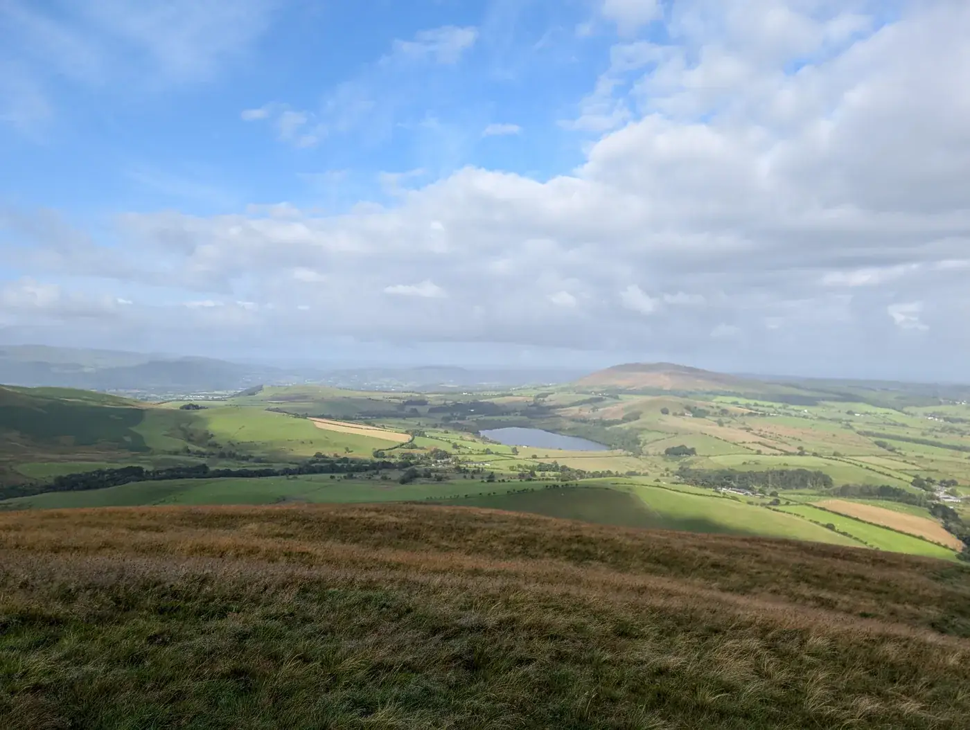 Longlands Fell - a grassy top above rolling green farmland, a small tarn and a rounded fell beyond under blue sky