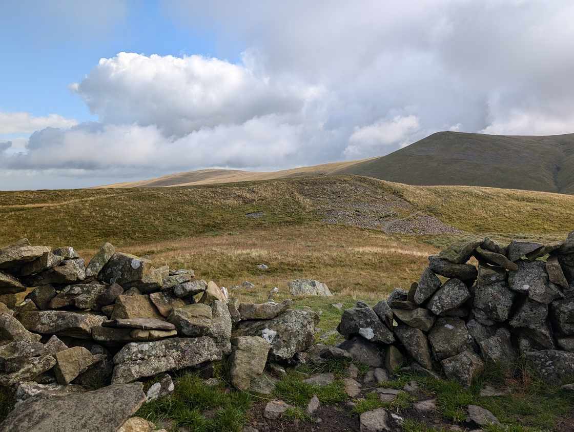Meal Fell - a broken stone shelter on a grassy top, broad grassy fells rising beyond under big clouds