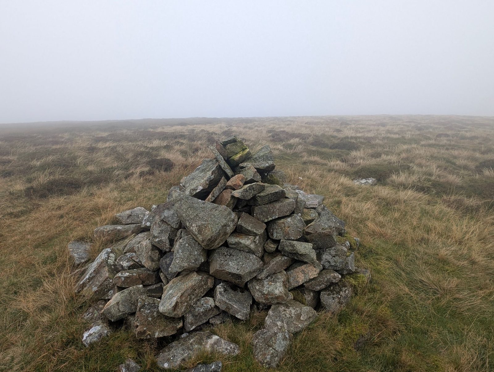 The summit cairn of Miller Moss, a pile of grey stones on a flat top of rough grass and heather, with mist hiding everything beyond.