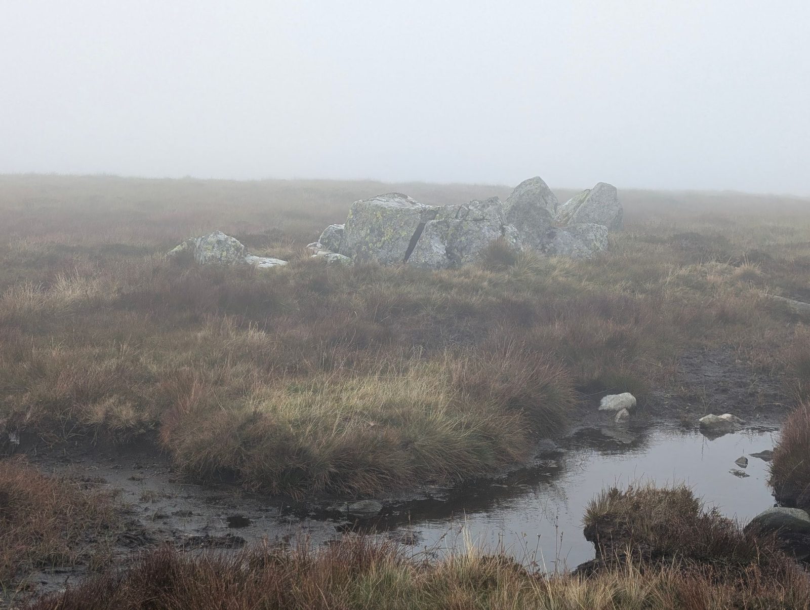 A cluster of grey boulders on the boggy moor of Miton Hill, with a peaty pool in the foreground and mist all around.