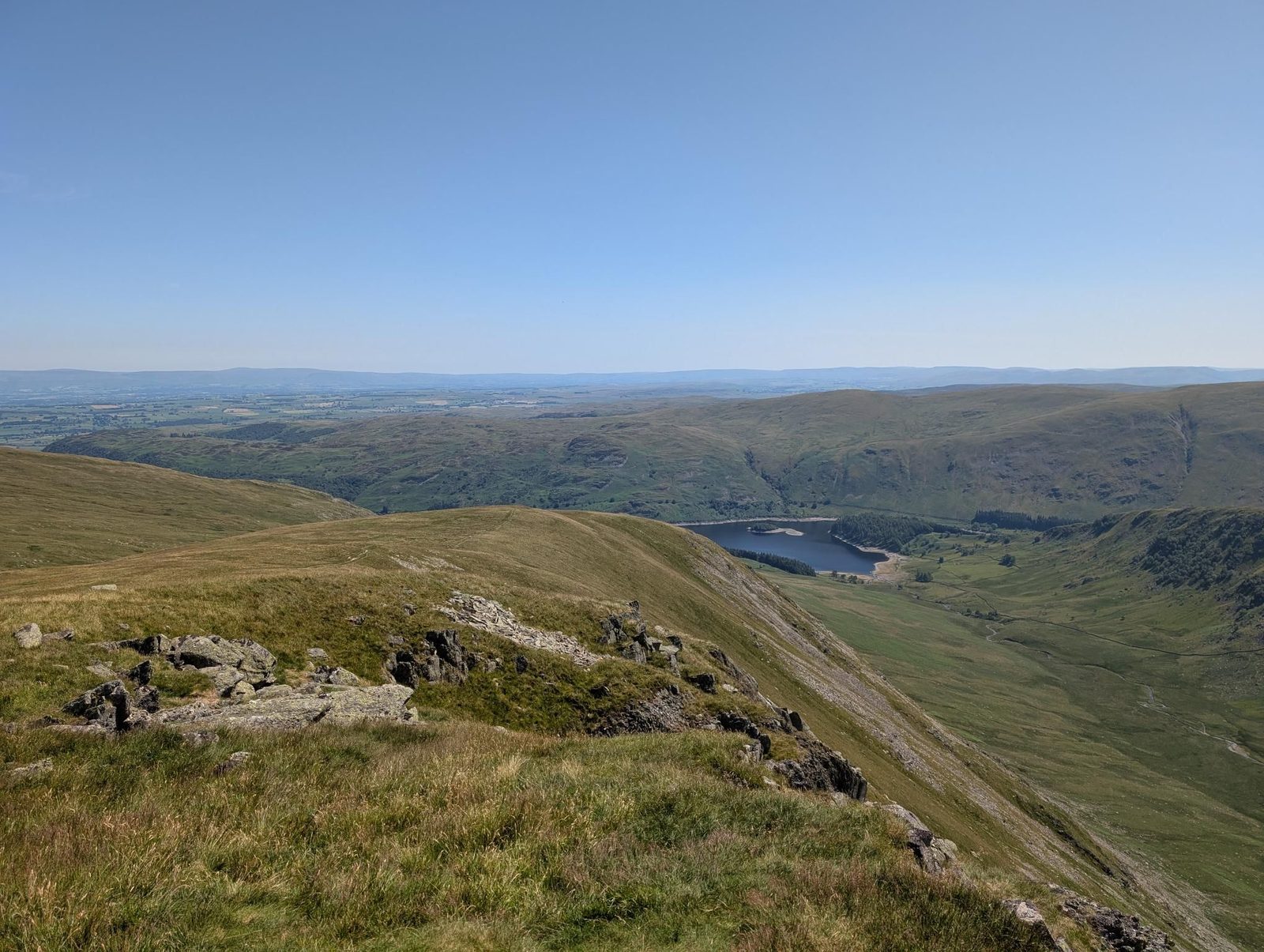 Looking down a steep grassy edge with rock outcrops into a valley with a reservoir, farmland stretching to the horizon under a clear sky.