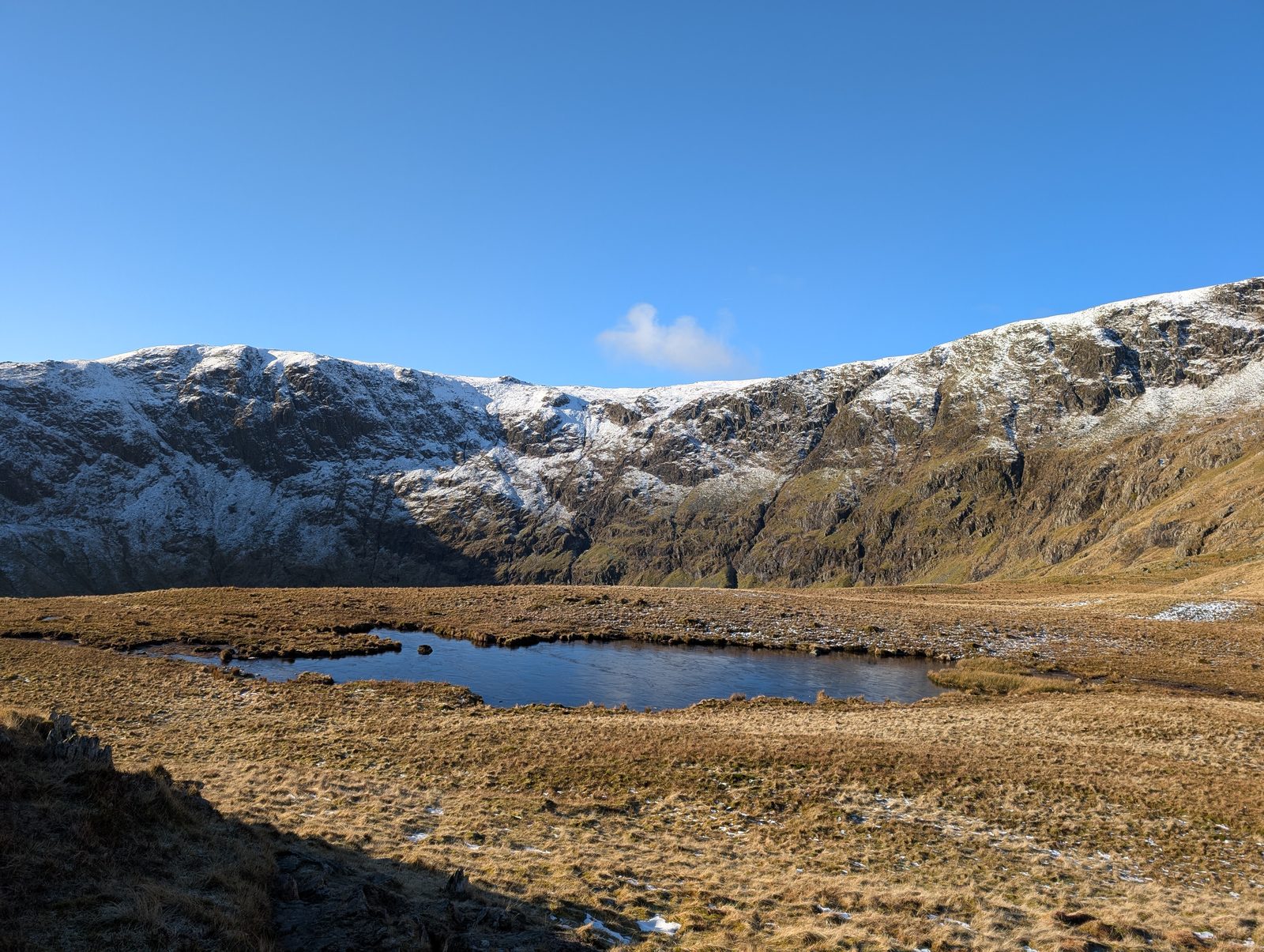A dark tarn below a long, snow-dusted line of crags, with russet grass in the foreground under bright winter sun