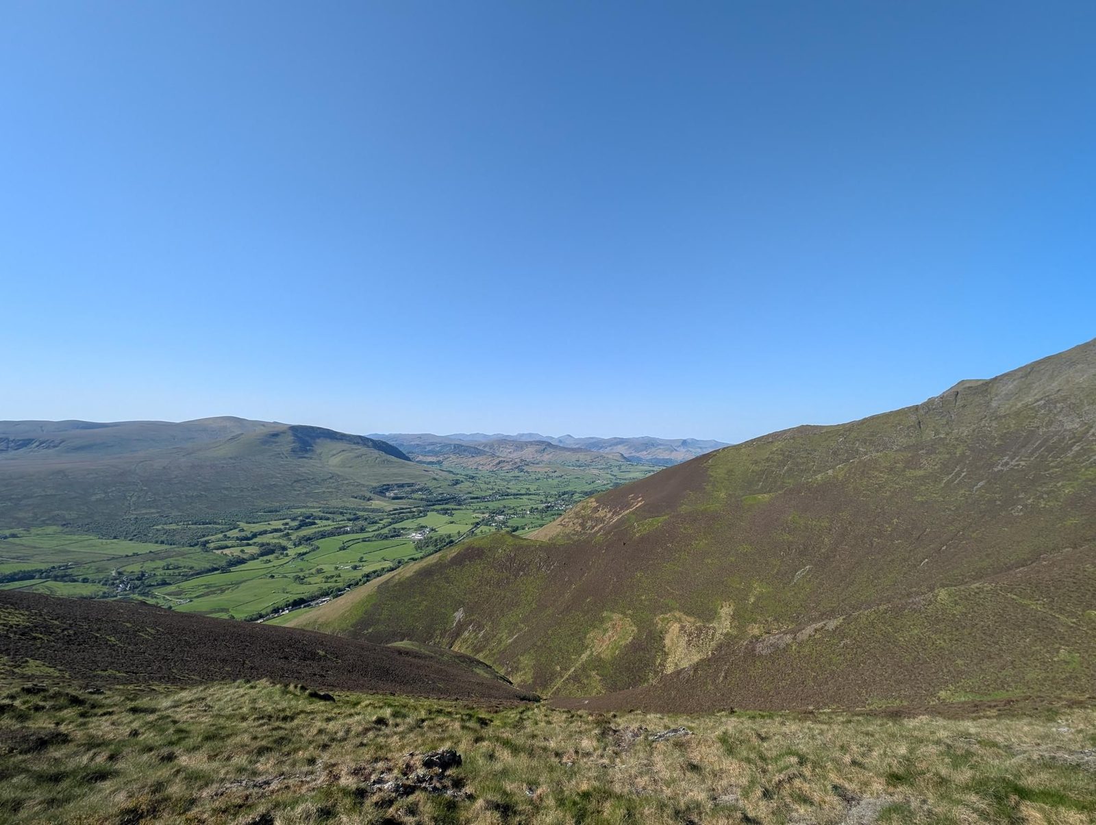 A steep heather slope dropping into a deep side valley, with green fields along the valley floor below and rounded fells under a cloudless blue sky