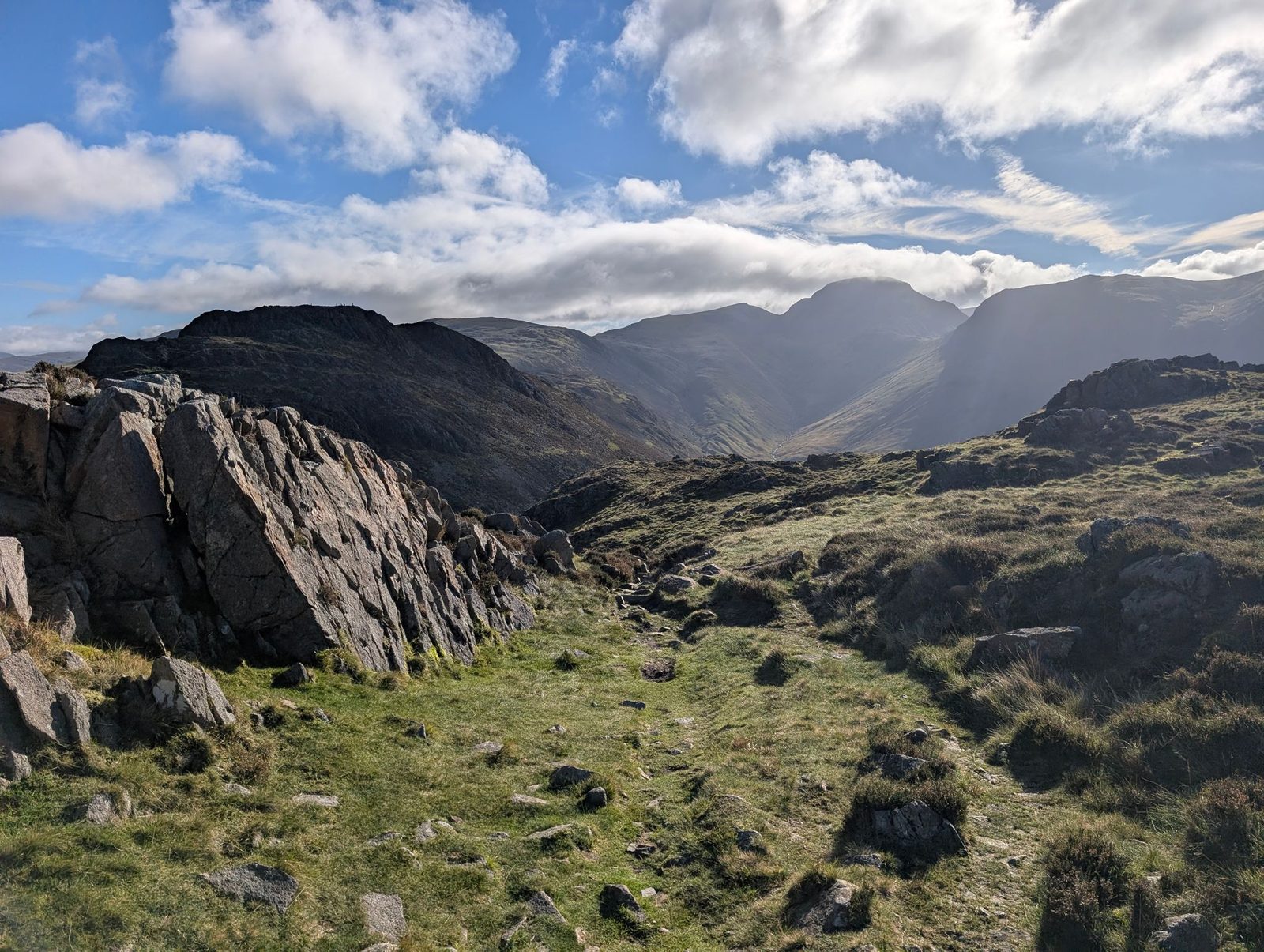 A grassy path between grey rock outcrops on Seat's top, looking across to steep high fells around a valley head under bright, broken cloud.