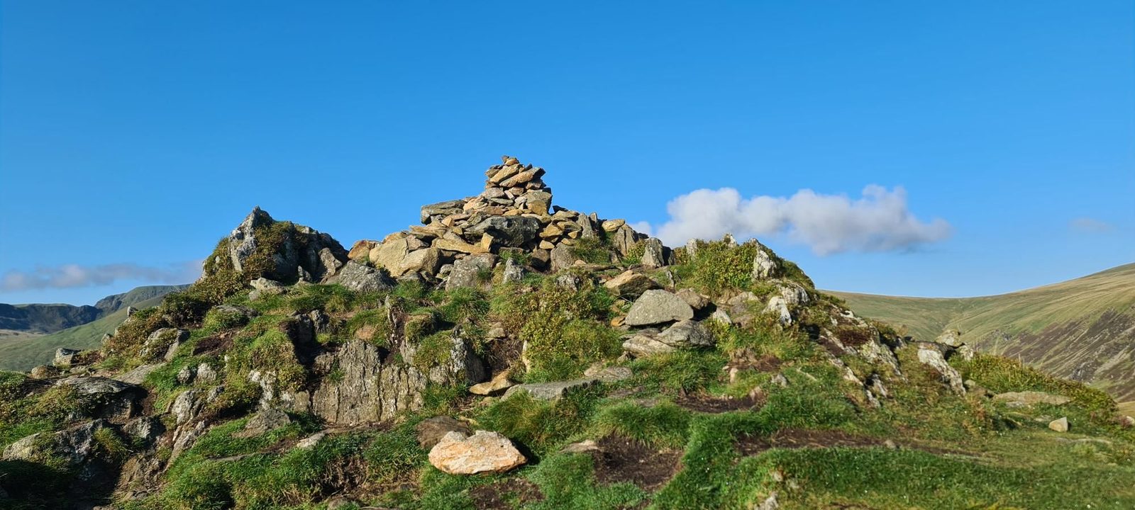 The stone cairn on Sheffield Pike's rocky summit knoll of grass and boulders, under a clear blue sky with grassy fells beyond.
