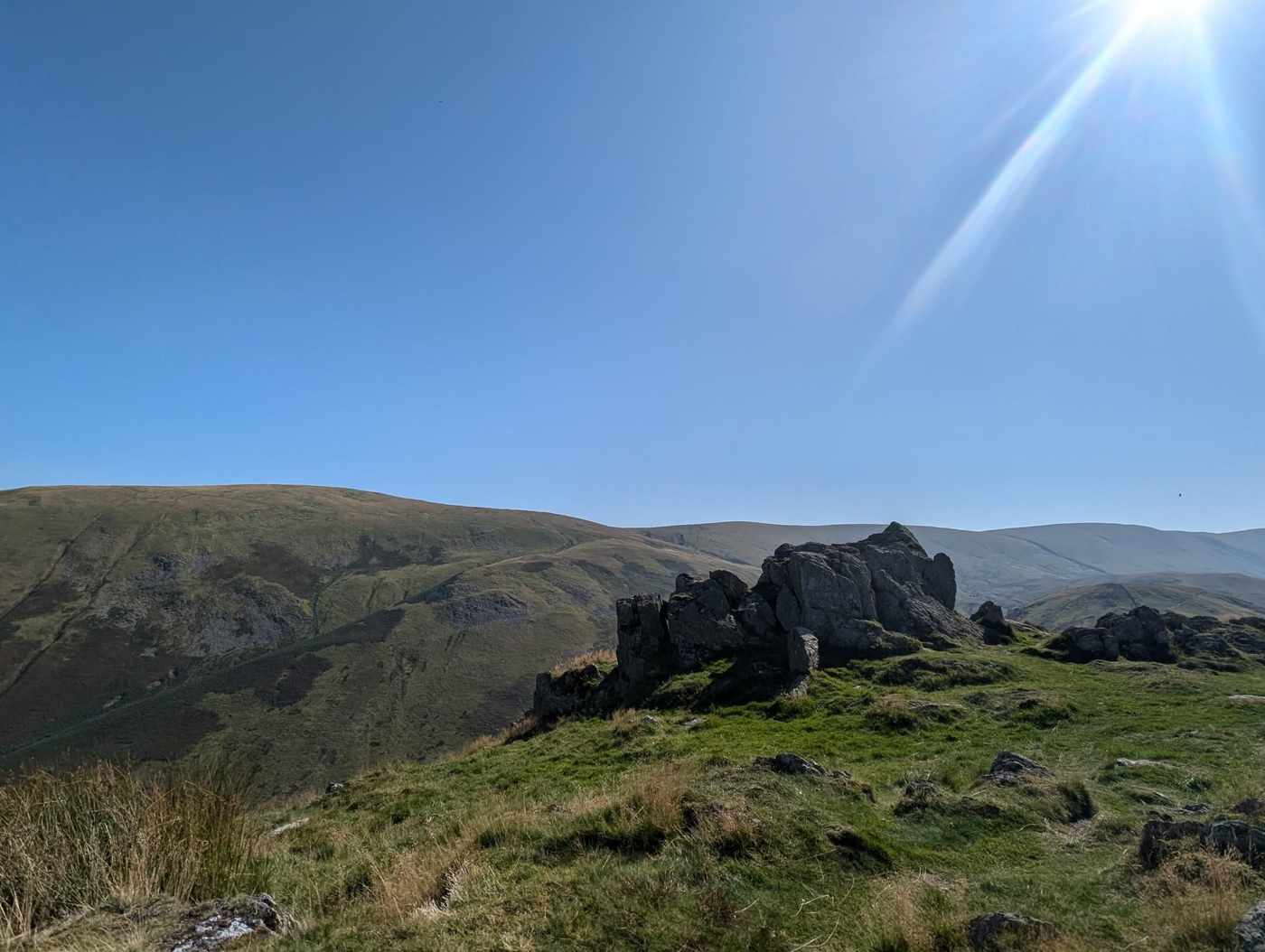Steel Knotts - the craggy summit tor of Pikeawassa on a grassy top, grassy fells beyond and the sun flaring above
