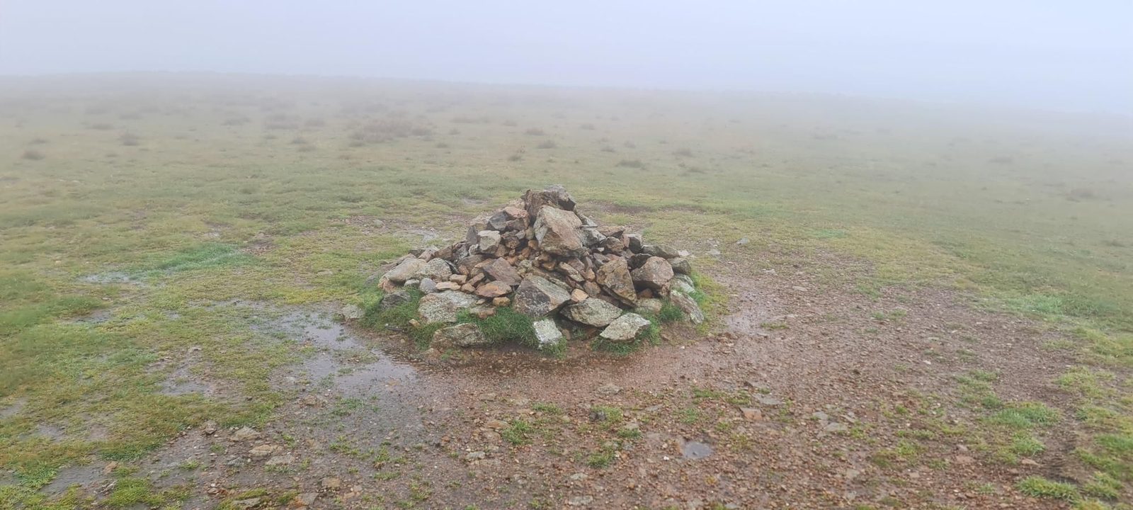 A low cairn of stones on Stybarrow Dodd's broad, wet grassy top, with puddles around it and mist hiding everything beyond.