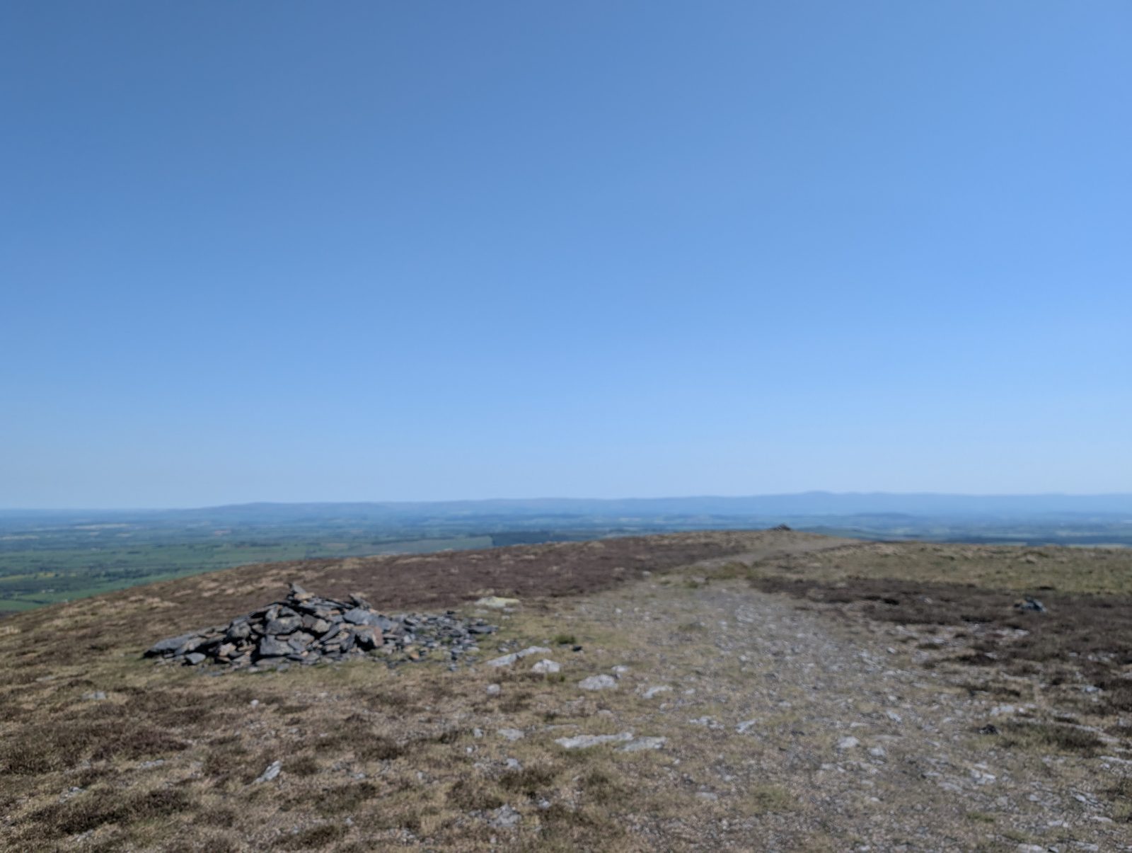 A small cairn on a stony, grassy ridge, with the lowlands stretching to the horizon under a clear sky