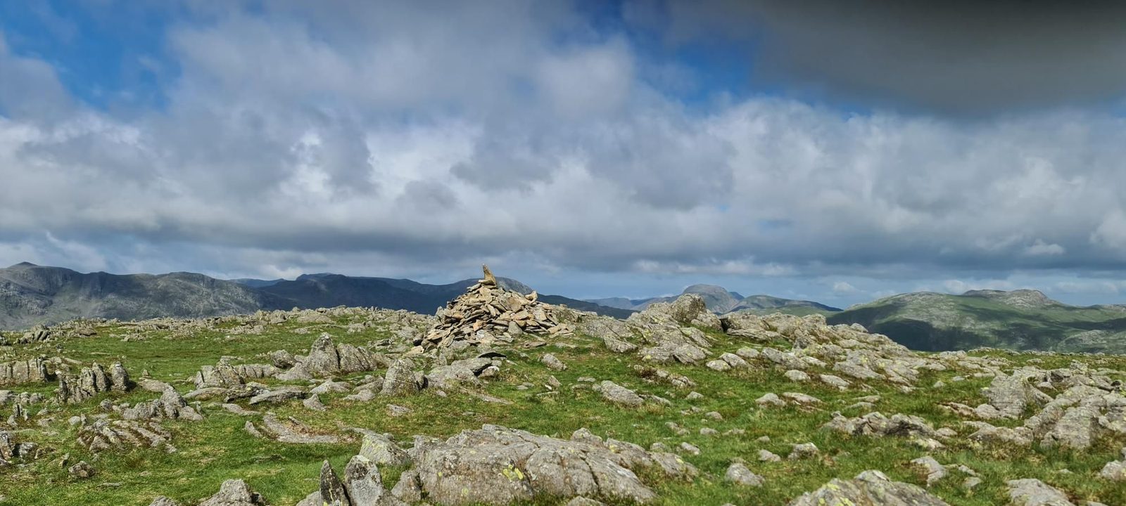 A stone cairn on Thunacar Knott's grassy top, strewn with grey rocks, with a line of distant fells under heavy cloud.