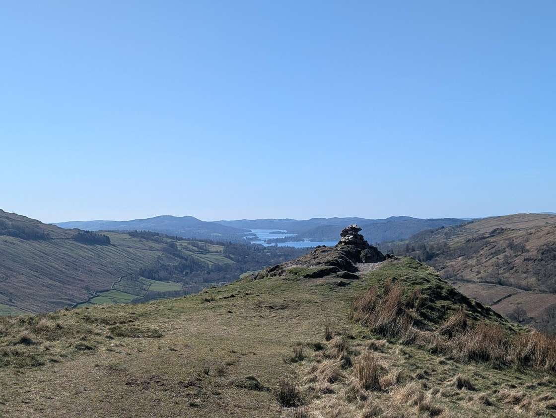 Troutbeck Tongue - a grassy knoll with a small cairn looking down the valley to Windermere shining under clear blue sky