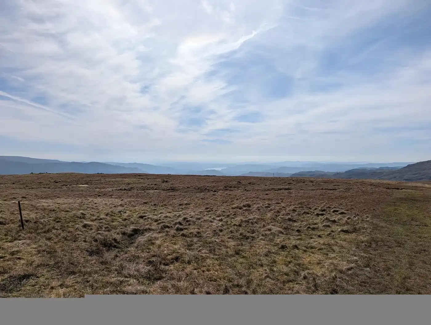 Ullscarf - a broad, flat moorland of rough brown grass with an old fence post, distant hazy fells beyond under high cloud