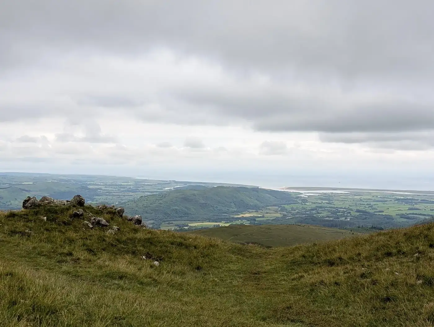 Whin Rigg - a grassy top above green coastal lowlands, the sea stretching along the horizon under grey cloud