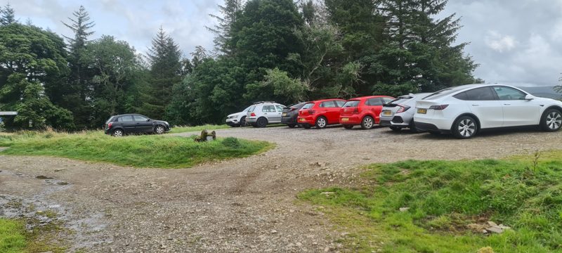 Gravel parking area at Low How on an August afternoon, full of cars, with woodland behind