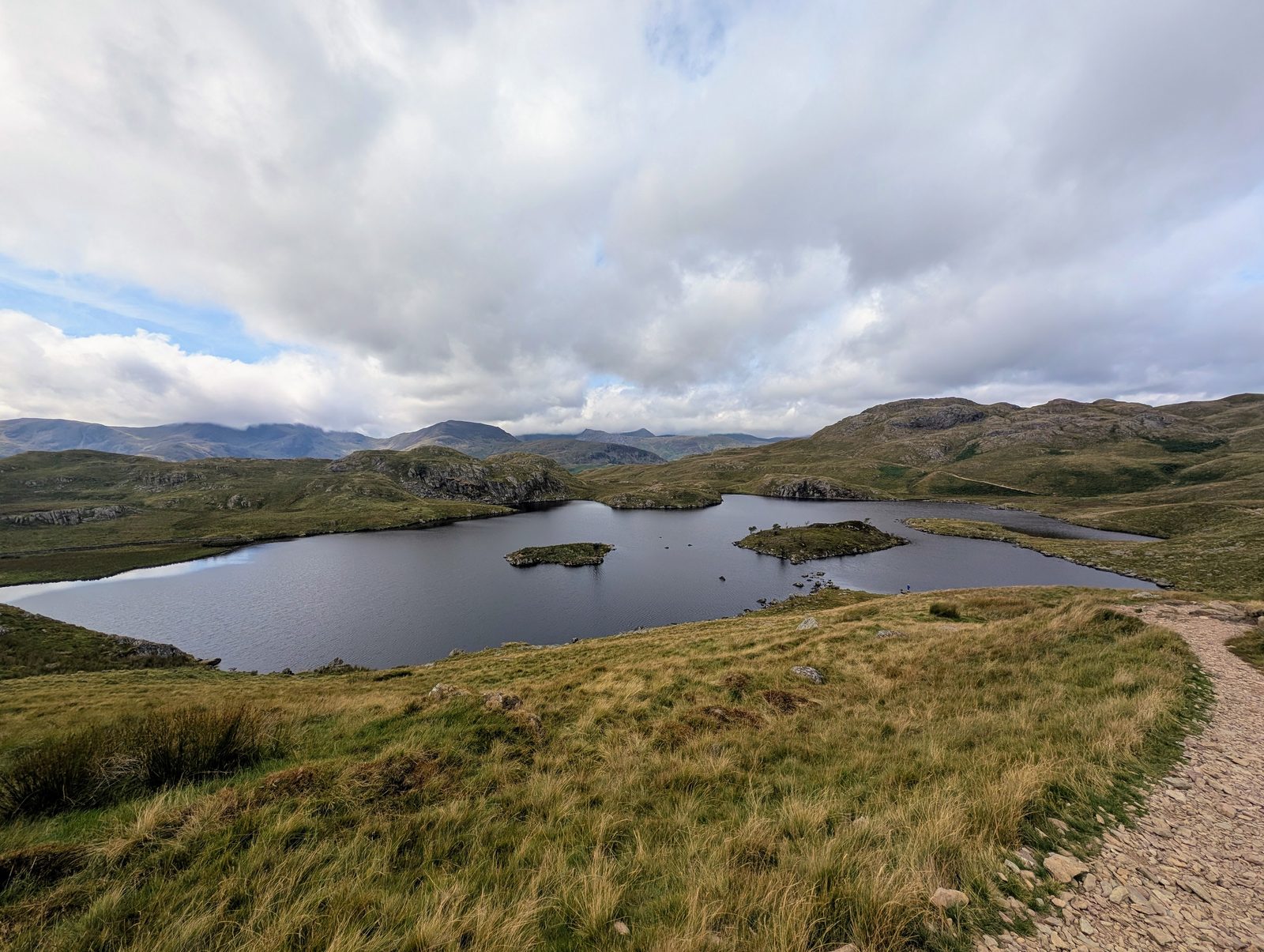 Angle Tarn from the path above it, two small grassy islets in the water, rough fells around it and distant hills under a cloudy sky