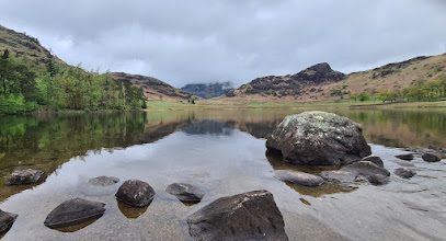 Boulders in the still shallows of Blea Tarn, a wooded shore on the left and the rocky Langdale Pikes on the skyline under grey cloud