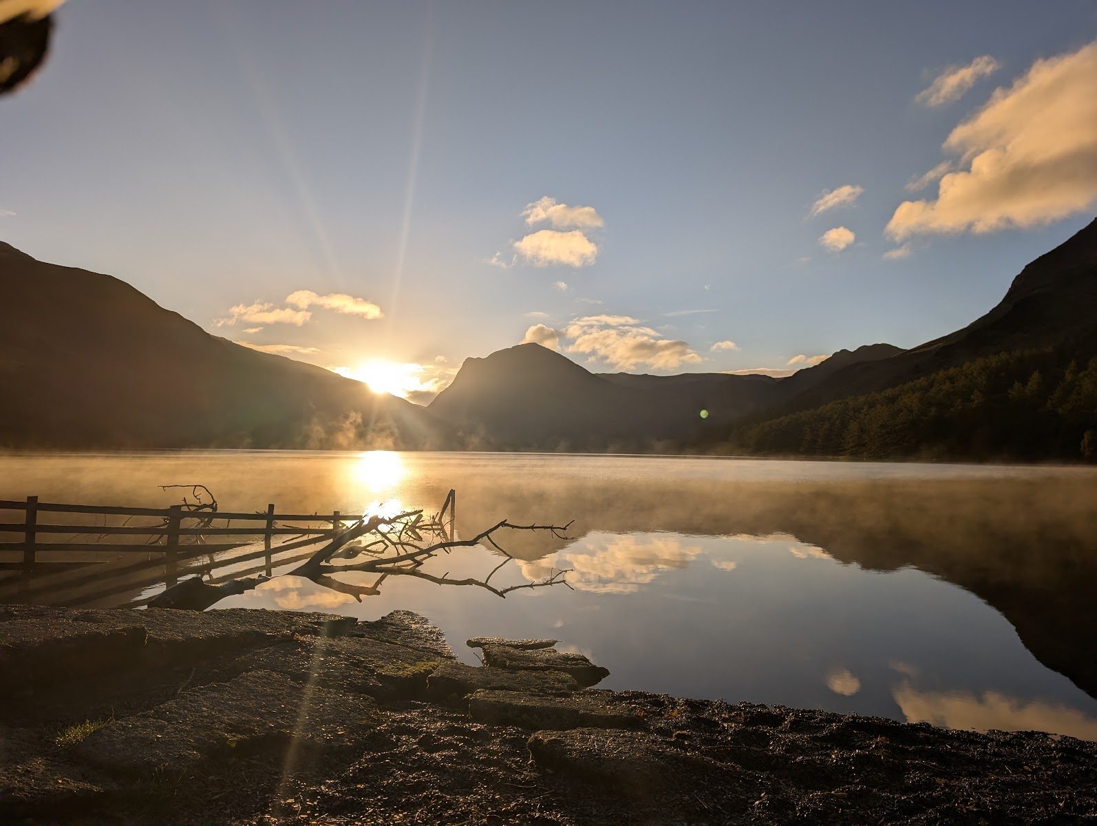 The sun low over the fells at Buttermere, mist rising off the still lake, an old fence and a fallen branch in the shallows, clouds mirrored in the water