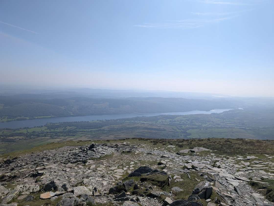 Coniston Water seen from a rocky fell top, the long lake stretching across the green valley below toward the hazy coast