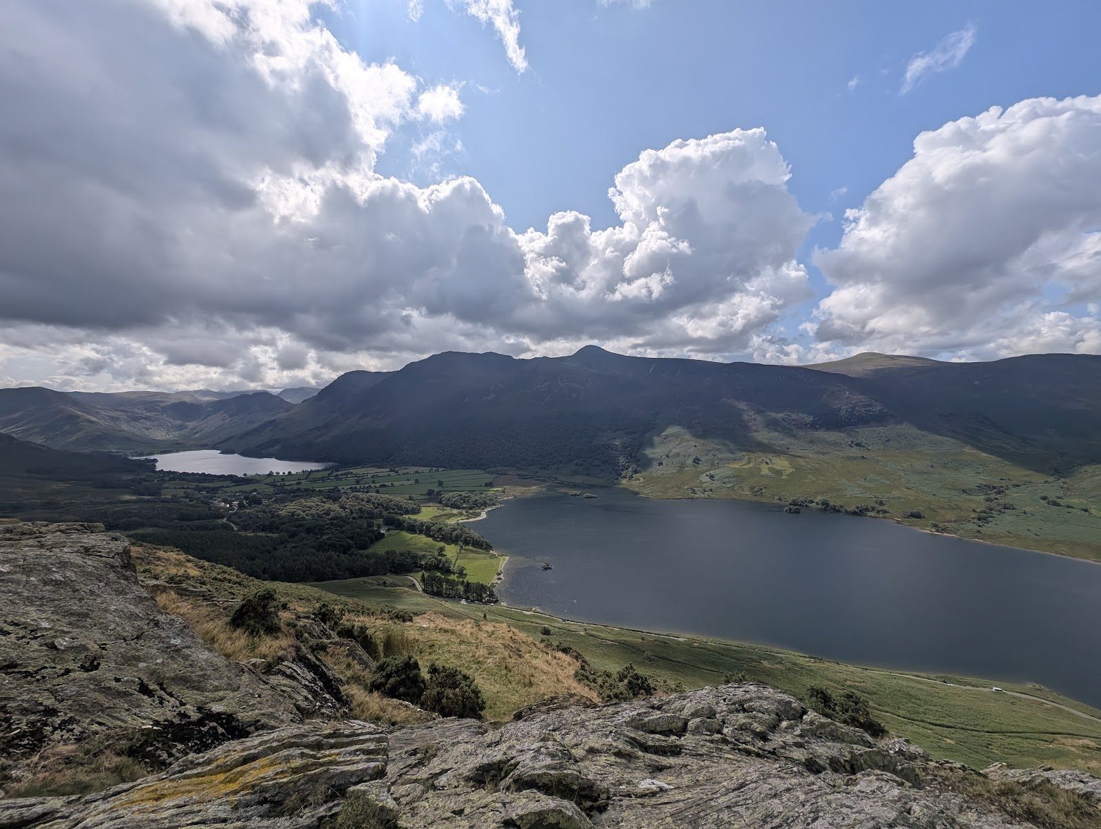 Crummock Water from a rocky outcrop high above its shore, Buttermere beyond the green fields between the two lakes, and a long dark ridge of fells under towering clouds